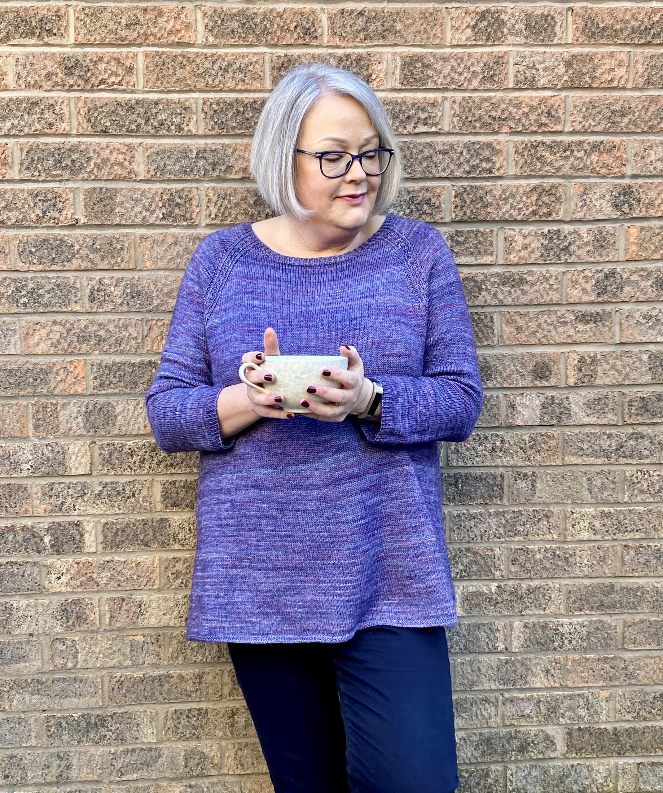 A women wearing a pale purple handknit sweater and holding a coffee cup leans against a wall looking towards the right of the picture