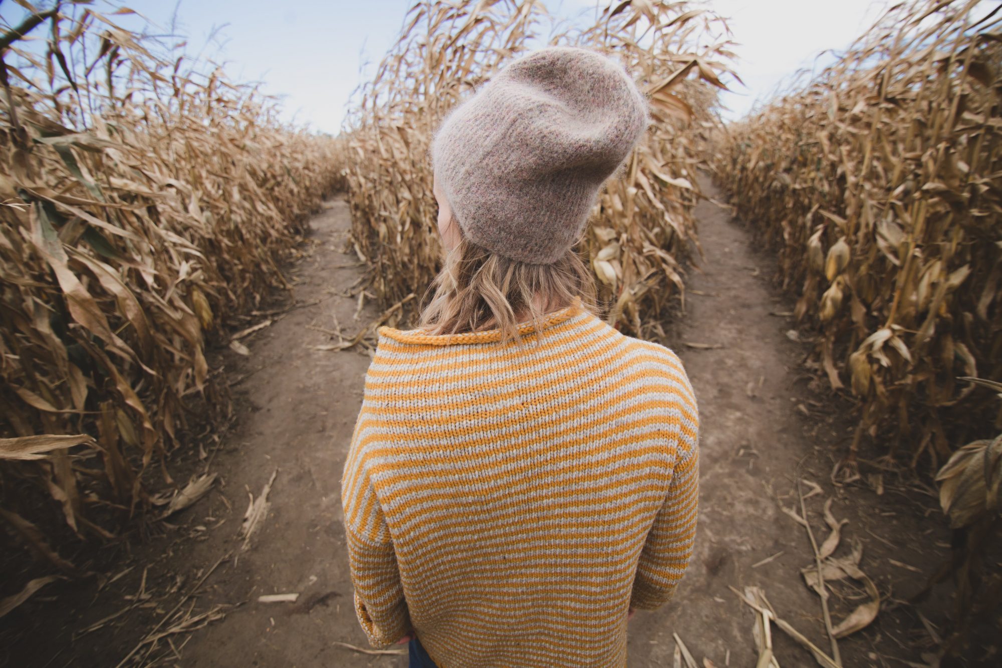 A woman in a yellow striped sweater and a grey hat stands looking at two pathways in a maze.