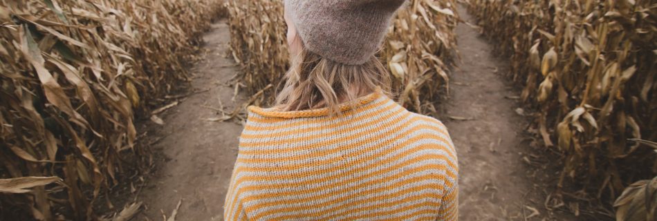 A woman in a yellow striped sweater and a grey hat stands looking at two pathways in a maze.
