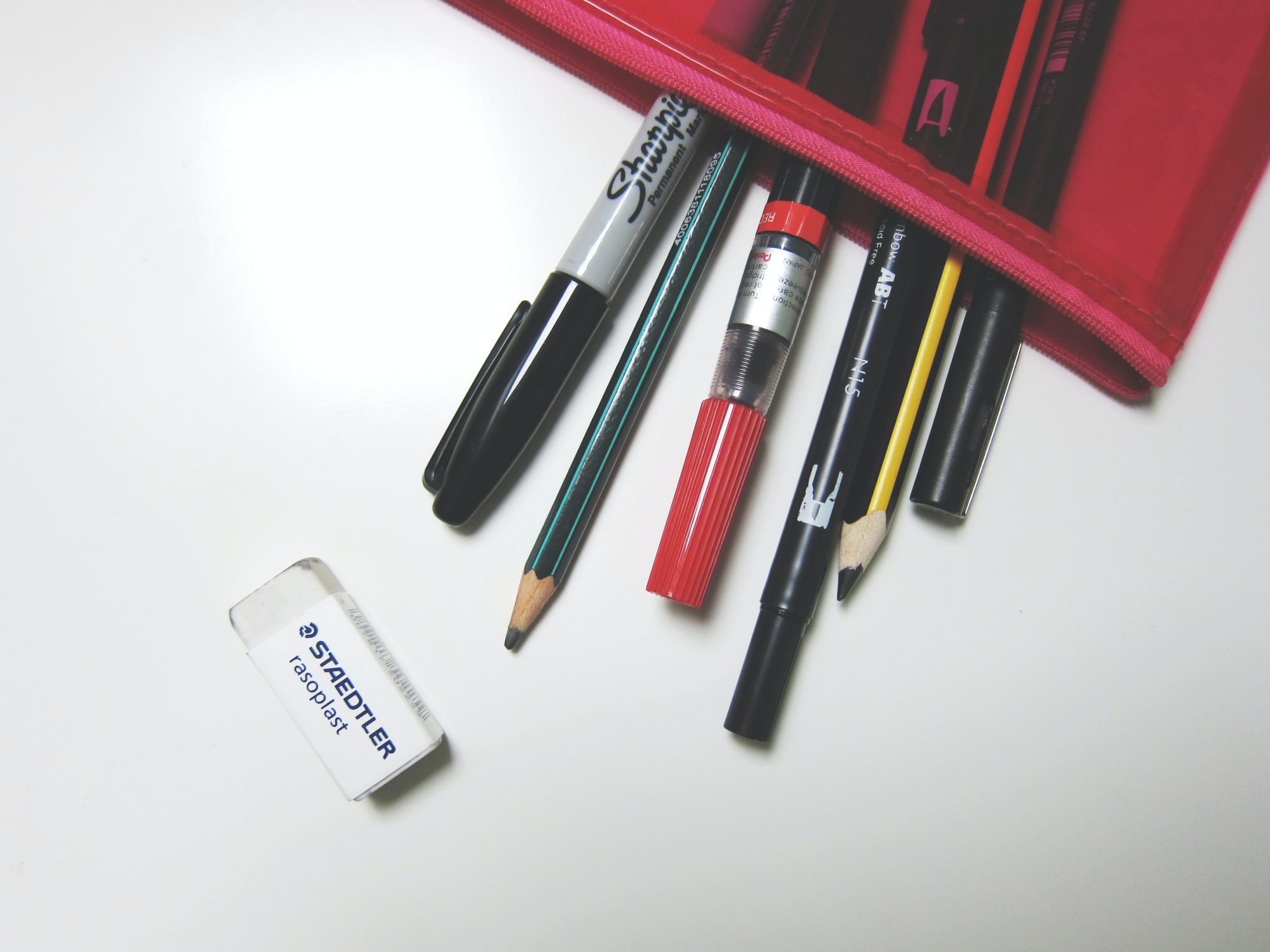 Picture shows a part of a red and black pencil case lying on a white surface with the zip open and pens and pencils spilling out. A white eraser lies next to them.