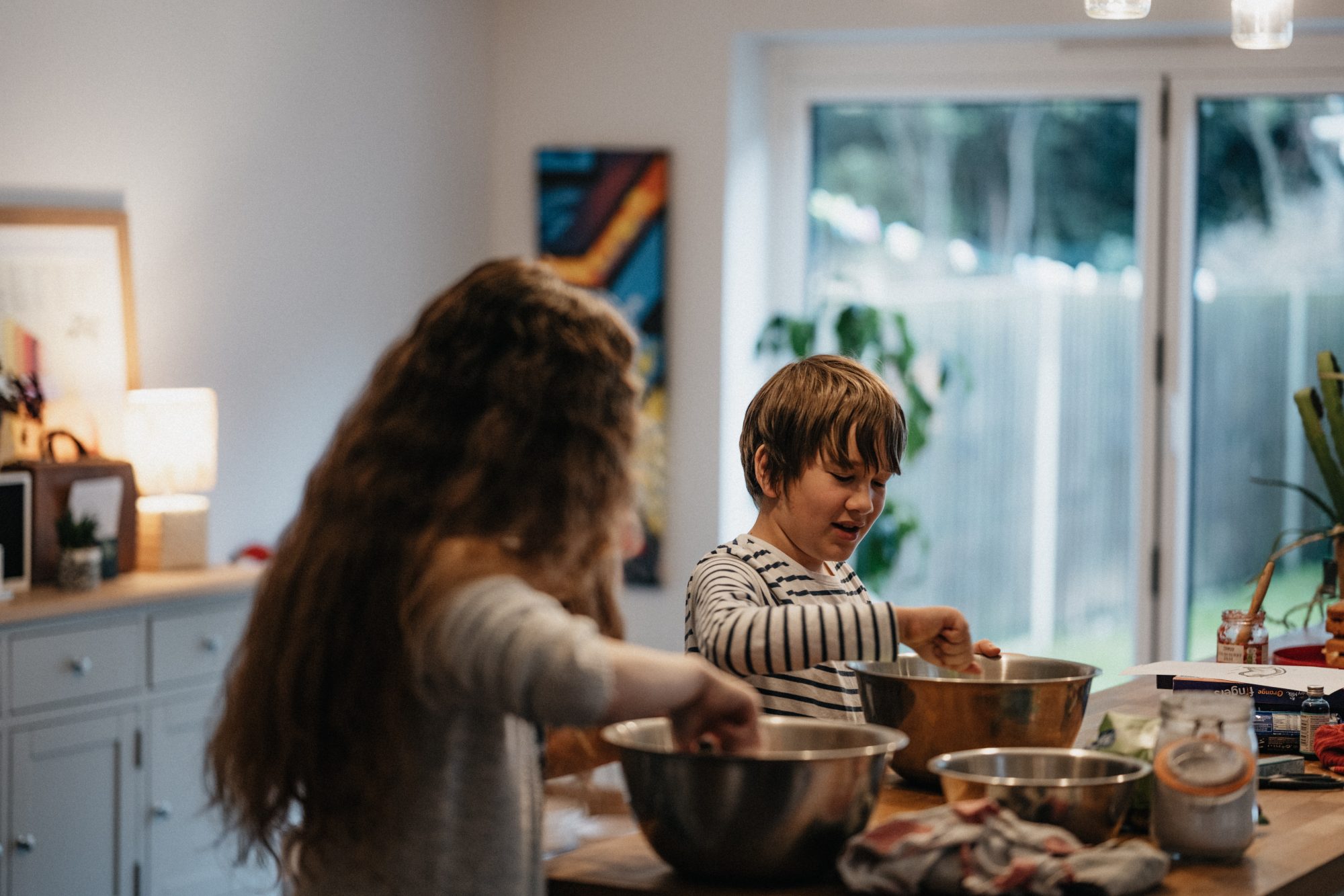 Two children, a boy and a girl, are stood in a light, airy kitchen at a wood work surface. They are each putting ingredients into steel mixing bowls. Other ingredients, books and bowls stand on the counter.They are baking their favourite treats to help them manage ADHD meds weight loss.