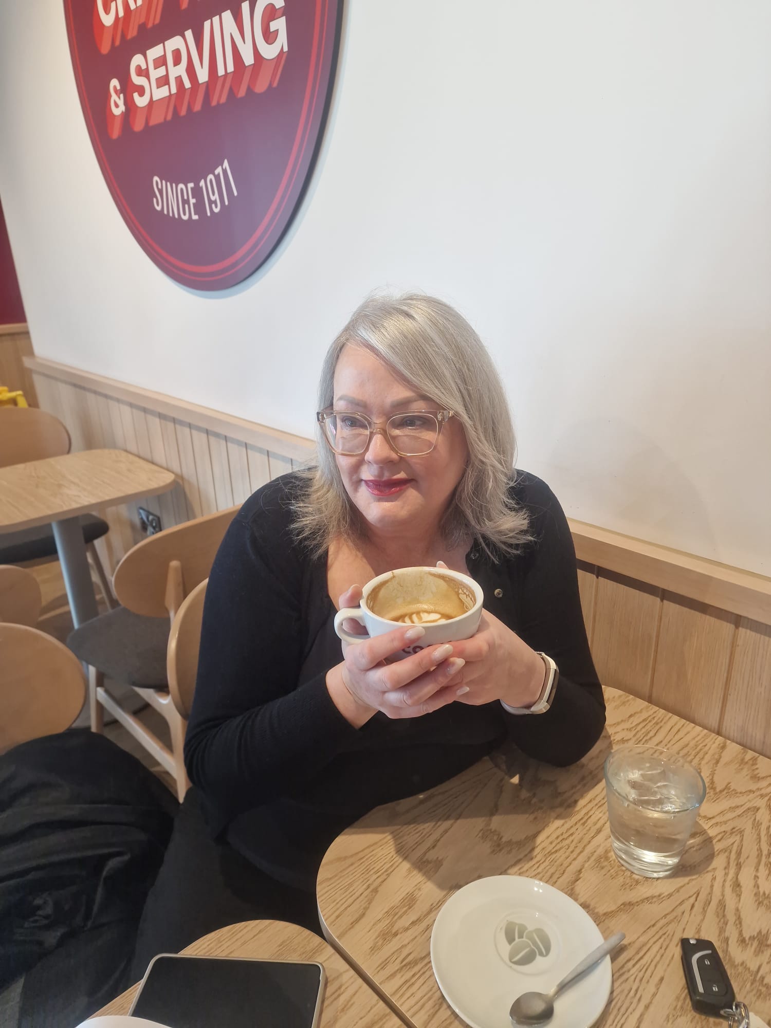 A white middle aged woman with silver hair and glasses sits at a modern wooden cafe table holding a cup of coffee in both hands. She is looking to her right and smiling gently.